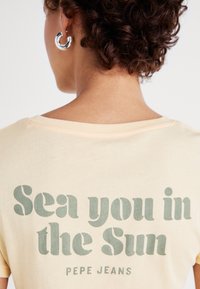 Žluté tričko s krátkým rukávem s zeleným nápisem "Sea you in the Sun" a "Pepe Jeans." Měkký bavlněný materiál, volný střih, kulatý výstřih.