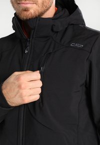 Veste isolée noire avec une poche poitrine zippée, logo sur l'épaule, doublure intérieure douce et capuche ajustable. Tissu lisse et texturé.