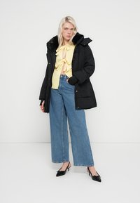 Svart dunjakke med pelskantet hette, lag på en gul knyttet topp og blå vidbente jeans, med svarte hælsko.