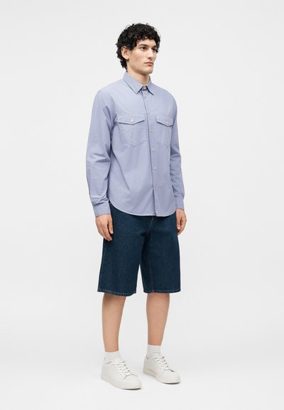 Νέος άνδρας όρθιος, φοράει ένα ανοιχτό μπλε πουκάμισο με τσέπες με καπάκια, σκούρες τζιν βερμούδες μέχρι το γόνατο, λευκές κάλτσες και λευκά αθλητικά παπούτσια.