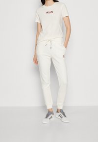 Kvinna som bär en vit Armani-t-shirt och matchande joggers med sneakers, stående framför en slät vit bakgrund.