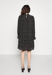 Svart klänning med vita prickar, långa ärmar, plisserad detalj vid midjan och en nyckelhålsöppning i ryggen, parat med svarta klackar.