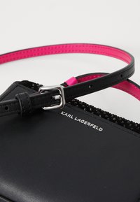 Črna usnjena torba z roza trakom, ki ima teksturiran črn detajl in srebrne dodatke. Logo "KARL LAGERFELD" v belem.