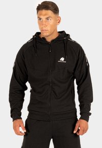 Zwarte zip-up hoodie van gestructureerde stof, met een logo op de borst en contrasterende gestreepte accenten langs de mouwen.