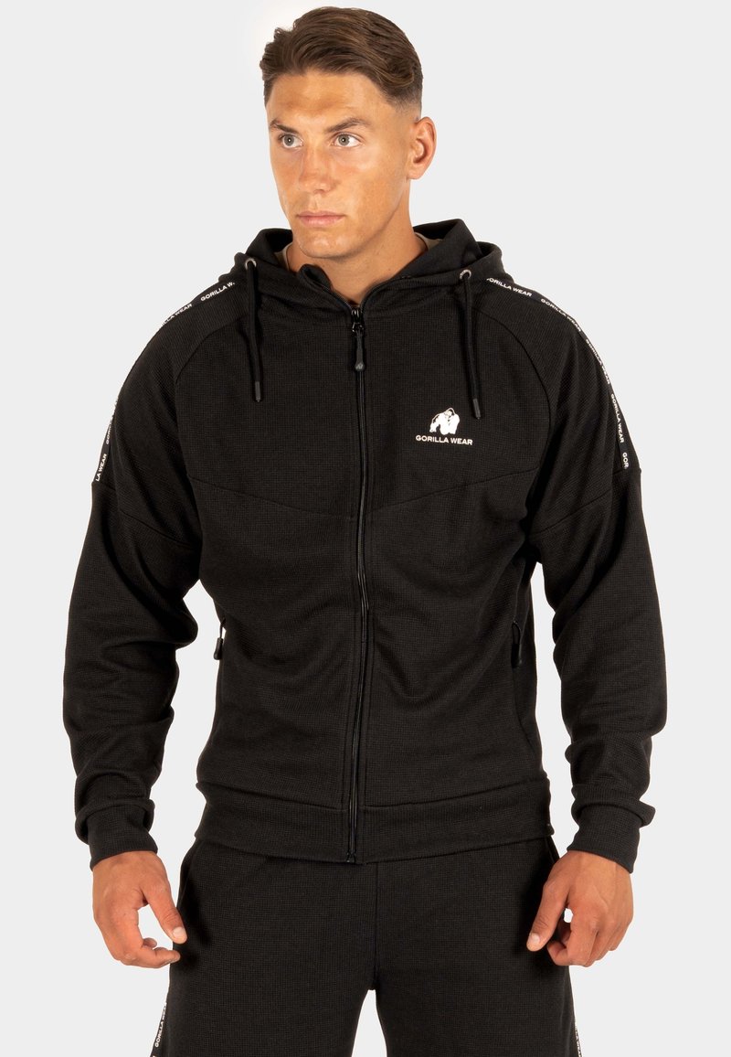 Zwarte zip-up hoodie van gestructureerde stof, met een logo op de borst en contrasterende gestreepte accenten langs de mouwen.
