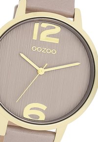 Montre ronde avec un boîtier en métal doré, cadran taupe texturé, chiffres dorés à 12 et 6 heures, et bracelet en cuir gris clair. Design minimaliste.
