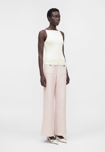 Γυναίκα που στέκεται φορώντας λευκό αμάνικο τοπ, φαρδιά ροζ ανοιχτό παντελόνια με μπροστινές τσέπες και λευκά μυτερά παπούτσια με λουράκια.