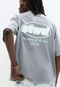 Grau T-Shirt mit kurzen Ärmeln, das das "Stwd"-Logo in mintgrün und den Text "Ich bin mit der Band unterwegs" auf dem Rücken zeigt. Lässige Passform, aus Baumwollstoff.