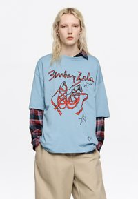 Camiseta de corte extragrande en azul claro con un gráfico de zapatillas de ballet rojas con cintas y texto manuscrito. Superpuesta a una camisa de franela a cuadros.