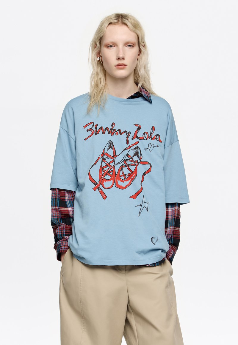 Camiseta de corte extragrande en azul claro con un gráfico de zapatillas de ballet rojas con cintas y texto manuscrito. Superpuesta a una camisa de franela a cuadros.