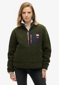 Pull en polaire vert avec une demi-zip, extérieur texturé, comportant une poche en tissu marine et un logo en patch. Jeans noirs portés.