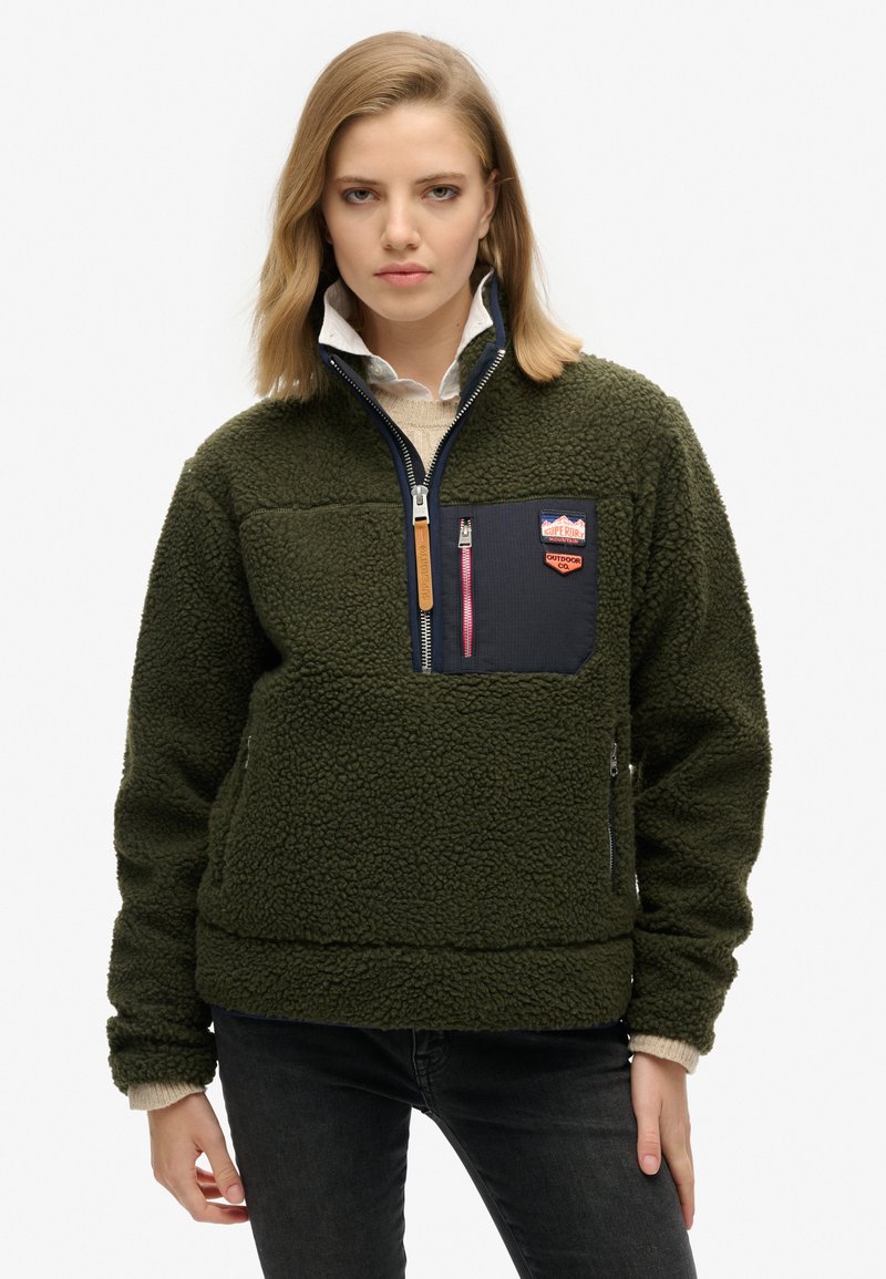 Pull en polaire vert avec une demi-zip, extérieur texturé, comportant une poche en tissu marine et un logo en patch. Jeans noirs portés.