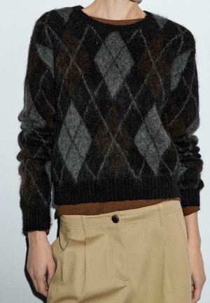 Personne portant un pull duveteux à motif losanges noir et gris sur une chemise marron, associé à un pantalon beige taille haute.