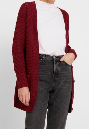Personne portant un cardigan bordeaux texturé par-dessus un t-shirt blanc et un jean noir délavé taille haute, une main dans la poche.
