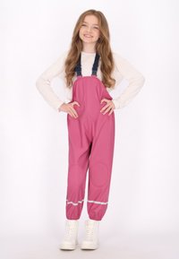 Ilgaplaukė mergina šypsosi, vilkėdama šviesią ilgomis rankovėmis marškinėlį ir rožinius neperšlampamus kombinezonus su atspindinčiomis juostelėmis bei baltas batus.