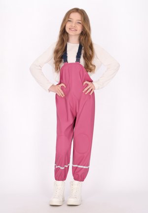 Flicka med långt hår som ler, iklädd en ljus långärmad tröja och rosa vattentäta overaller med reflexstrimmor och vita stövlar.