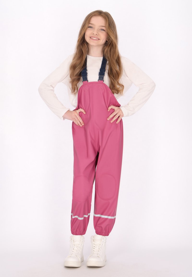 Ilgaplaukė mergina šypsosi, vilkėdama šviesią ilgomis rankovėmis marškinėlį ir rožinius neperšlampamus kombinezonus su atspindinčiomis juostelėmis bei baltas batus.