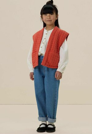 Een jong meisje staat met een witte blouse met knopen, een oranje gewatteerd vest, opgerolde blauwe jeans, witte sokken en zwarte schoenen tegen een effen achtergrond.