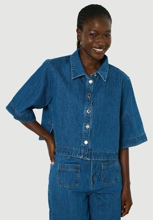 Vrouw die glimlacht, draagt een blauw jeansjack met fijne strepen en korte mouwen en bijpassende hoog getailleerde broek tegen een effen achtergrond.