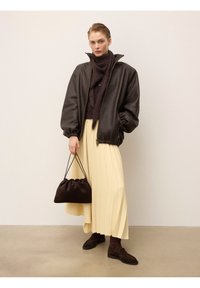 Veste bomber en cuir marron foncé, jupe plissée crème, écharpe en tricot marron et sac à main. Chaussures marron avec des accents texturés. Fond lisse.