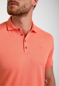 Koralno oranžna polo majica z teksturirano obliko, tremi rjavimi gumbi na ovratniku in majhnim vezenim logotipom na prsih.