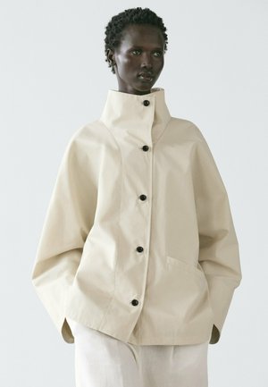 Model nosí volnou béžovou bundu s vysokým límcem, černými knoflíky a kapsami, doplněnou světle zbarvenými kalhotami, na jednobarevném pozadí.