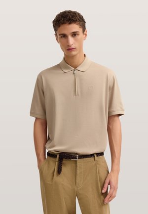 Junger Mann trägt ein beiges Polohemd mit kurzen Ärmeln und Reißverschluss sowie hellbraune Hose mit geflochtenem dunkelbraunem Gürtel.