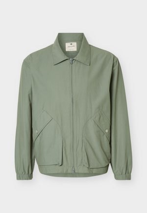 Giacca verde chiaro con zip, colletto appuntito, due tasche con patta e bottoni a pressione e polsini elasticizzati, mostrata su uno sfondo chiaro e semplice.
