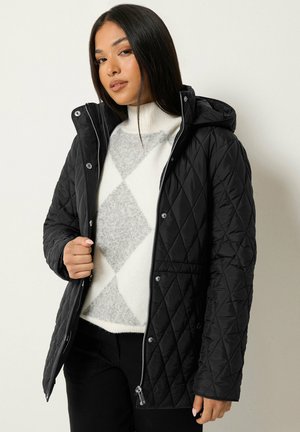 Femme portant une veste noire matelassée à capuche sur un pull blanc et gris à motif losange, debout devant un fond clair uni.