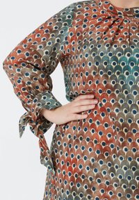 Kleurige blouse met patroon, poffige mouwen en strikaccenten. Heeft een herhaald ontwerp van rode, blauwe en bruine schubben op een lichte achtergrond.