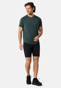 T-shirt sportivo verde scuro a maniche corte, abbinato a pantaloni corti neri attillati. L'outfit presenta dettagli riflettenti e scarpe da ginnastica nere.