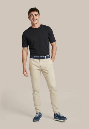 Jonge man die lacht, draagt een zwart shirt met korte mouwen, beige broek met blauwe riem en blauwe sneakers, staand tegen een effen achtergrond.