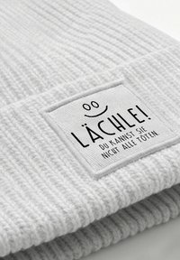 Folded white knitted fabric with a sewn square patch showing a smiley and German text "Lächle! Du kannst sie nicht alle töten."