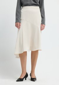 Een beige, knielange rok met een uitlopende zoom en diagonale naaddetails, gecombineerd met zwarte, puntige hakken. De stof lijkt glad en lichtgewicht.