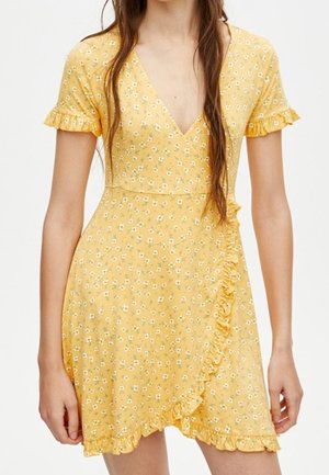 Robe portefeuille jaune à manches courtes avec un petit imprimé floral blanc et des bords à volants portée par une personne aux longs cheveux bruns.