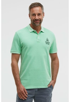 Lächelnder Mann, der ein hellgrünes Poloshirt mit dem Logo "Gaastra Nautical Supplies" auf der Brust und graue Hosen trägt, und mit einer Hand in der Hosentasche posiert.