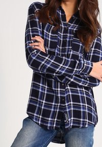 Woman wearing a navy, white, and blue plaid button-up shirt with long sleeves, paired with light blue jeans, arms crossed.