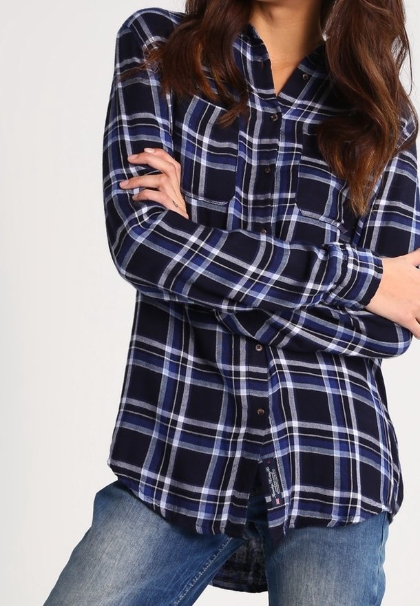 Woman wearing a navy, white, and blue plaid button-up shirt with long sleeves, paired with light blue jeans, arms crossed.