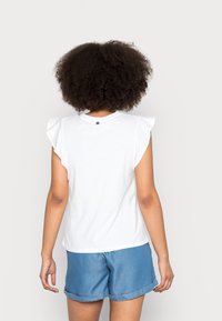 Camiseta blanca con mangas acampanadas, escote redondo y un pequeño acento dorado en la parte trasera. Combinada con pantalones cortos azules.