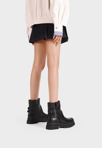 Stivali neri in pelle alla caviglia con suola spessa, cerniera laterale e due dettagli con fibbia. Abbinati a una gonna scura e una giacca chiara.