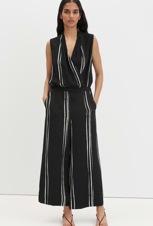 Femme portant une combinaison noire sans manches avec des rayures verticales blanches, debout les mains dans les poches devant un fond uni.