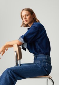 Denim overhemd in donkerblauw met opgerolde mouwen, en hooggewaisted donkerblauwe jeans. Model zit op een metalen stoel met houten accenten.