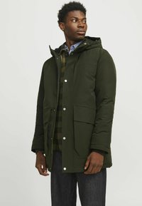 Roheline polsterdatud jakk, millel on kapuuts, nööpidega kinnitused, suured ees taskud ja sile tekstuur. Kandsen plaidi muster särgi ja tumedate pükstega.