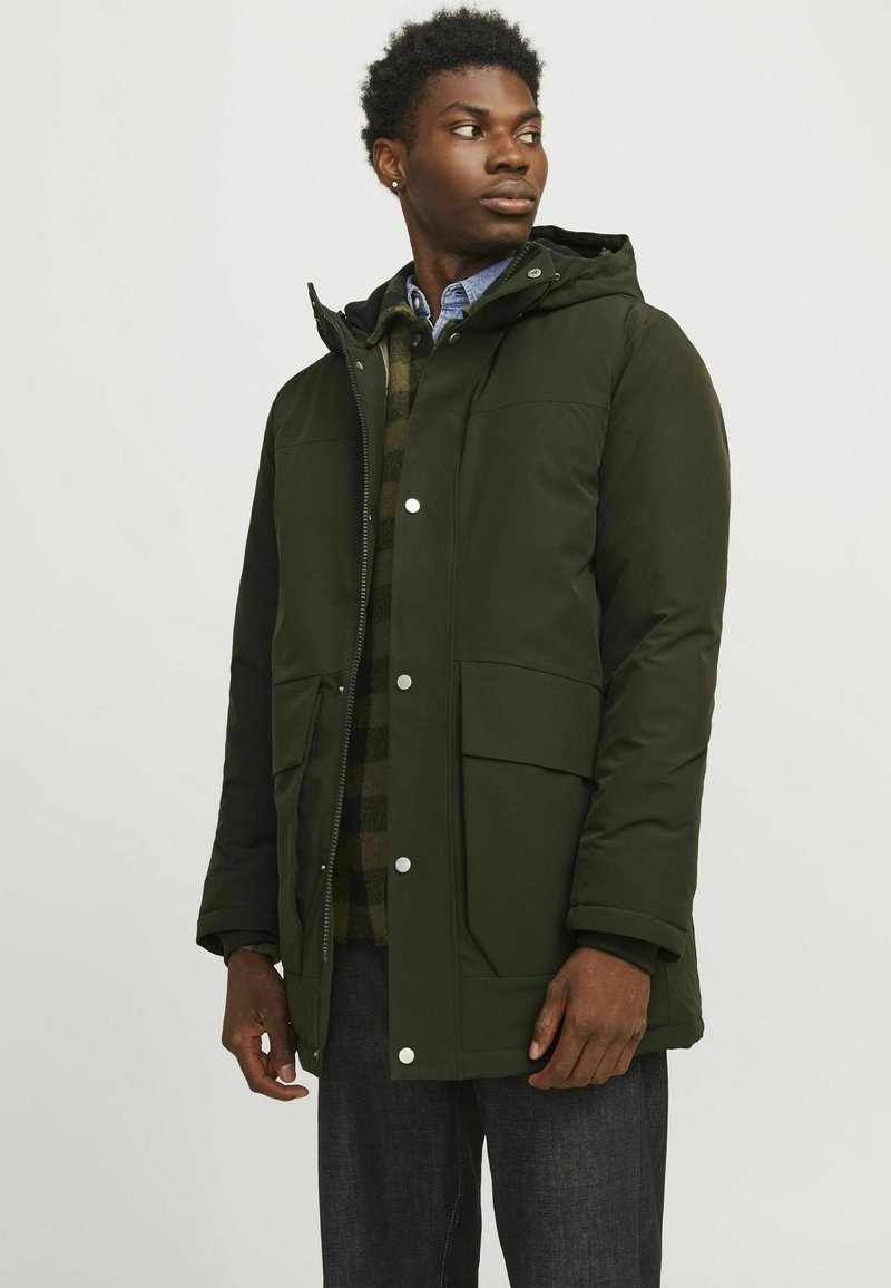 Roheline polsterdatud jakk, millel on kapuuts, nööpidega kinnitused, suured ees taskud ja sile tekstuur. Kandsen plaidi muster särgi ja tumedate pükstega.