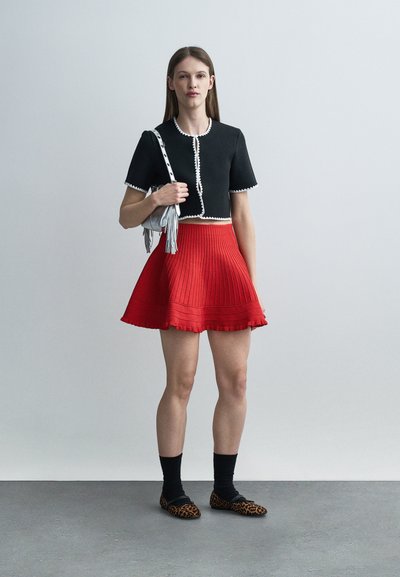 Γυναίκα με μαύρο κοντό τοπ με λευκή λεπτομέρεια, κόκκινη πλισέ φούστα, μαύρες κάλτσες, παπούτσια με μοτίβο λεοπάρ και κρατώντας ασημένια τσάντα πάνω σε γκρι δάπεδο.