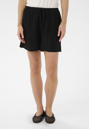 Kvinde iført løse sorte shorts med snore, hvid top, guldbøjler og sorte flade sko i mesh, stående på hvid baggrund.