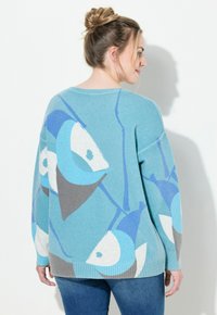 Ljusblå tröja med fiskmönster i blå och grå nyanser. Har rund halsringning och ribbade ärmslut. Mjuk stickad textur.