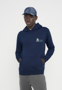 Μπλε ναυτικό φούτερ με τσέπη καγκουρό και γραφική εκτύπωση που γράφει «Cinéma Surréaliste». Συνδυασμένο με γκρι φόρμα και καπέλο με σχέδιο.