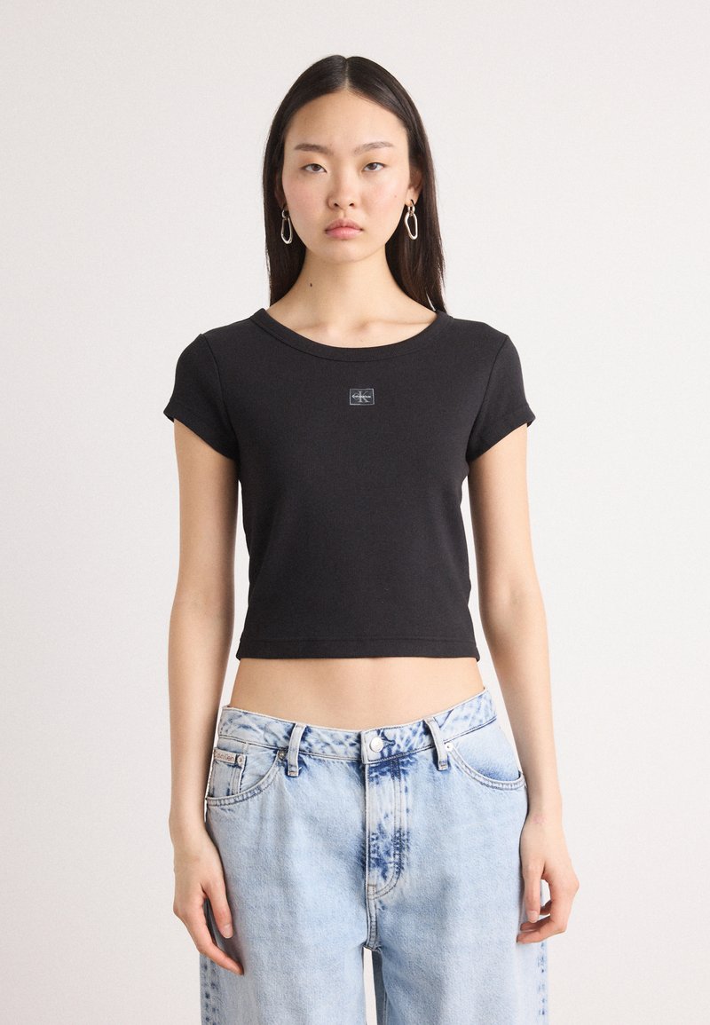 Black cropped t-shirt made of cotton with a round neckline and short sleeves, featuring a small logo on the front. Paired with light blue jeans.