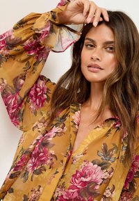 Blumige Bluse in senfgelb mit langen, durchsichtigen Ärmeln. Verfügt über große rosa und bordeauxrote Blumenmuster sowie einen V-Ausschnitt.